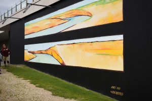 Man gesturing at a mural. Mural is of two rectangles painted horizontally on a black backgrpund. If features wattery areas of blue, yellow and orange which look like a landscape.