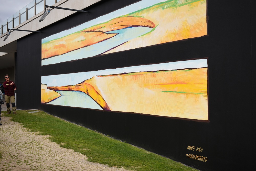 Man gesturing at a mural. Mural is of two rectangles painted horizontally on a black backgrpund.  If features wattery areas of blue, yellow and orange which look like a landscape.