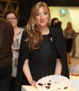 Woman in black cape with long red hair smiles at camera in front of artwork.