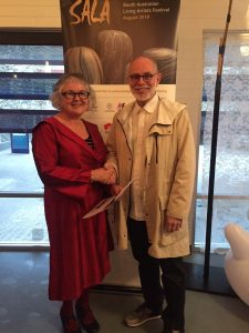 Woman in read dress smiles holding a certificate in one hand and with the other shaking hands with an older gentleman. both are smiling and looking at the camera