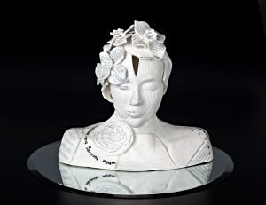 A white ceramic bust of a person sitting on a round mirror with a black background. There are textural patterns on the skin and flowers and leaves in the hair.