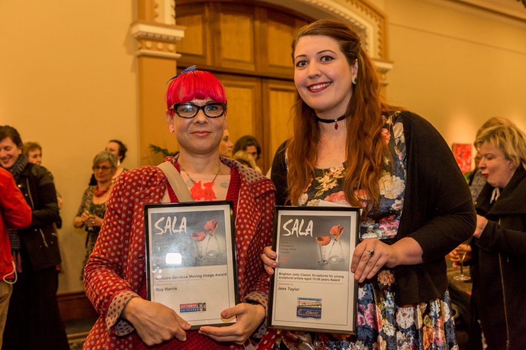 Two women standing next to eachother holding framed awards. The one on the left has pink hair and the one on the right has long brown hair. 