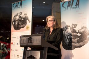 Woman stands in front of SALA banners and speaks, smiling into a microphone. 