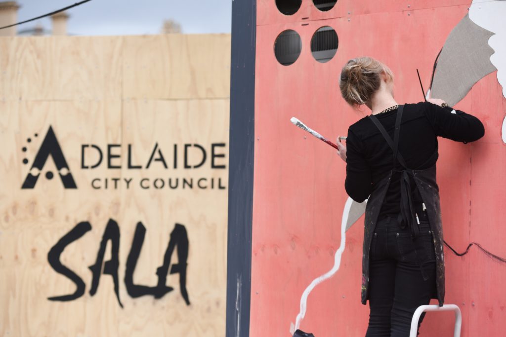 Female artist standing on a ladder painting on board with pink grey and white paint. 