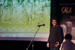 man standing at a microphine with a projected image in the background. He is gesticulating to an unseen crowd and smiling.
