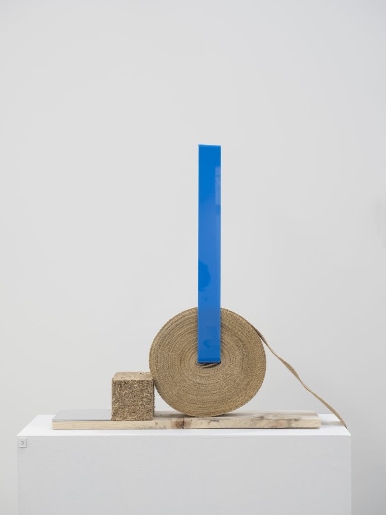 A roll of hesshan looking ribbon sits on a plinth with a rectangular mid blue shape protruding from the middle. On the left of the roll a wooden cube sits touching the roll. 